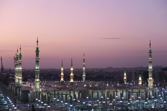 The lit minarets and courtyard of Masjid an-Nabawi in Madinah under a pink and violet dusk sky.