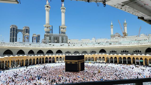 The Kaaba surrounded by pilgrims in the courtyard of Masjid al-Haram, with the arcades and minarets of the mosque under a blue sky.