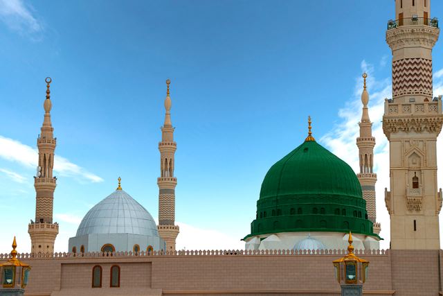 The Green Dome and silver dome of Masjid an-Nabawi in Madinah with minarets against a blue sky.