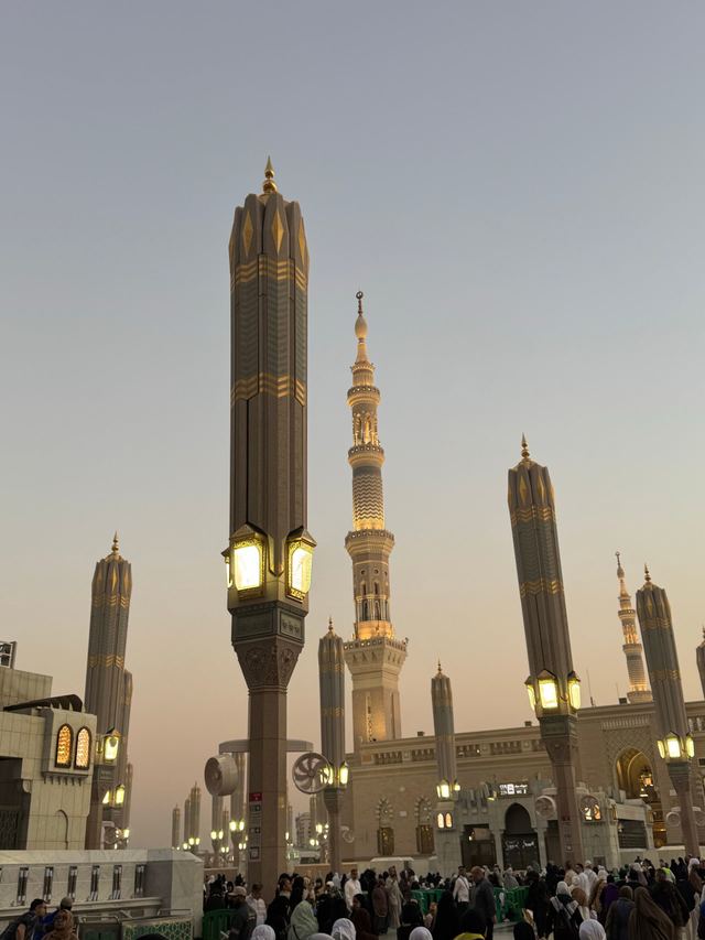 Folded umbrella pillars with lanterns in the courtyard of Masjid an-Nabawi in the early evening, minarets behind.